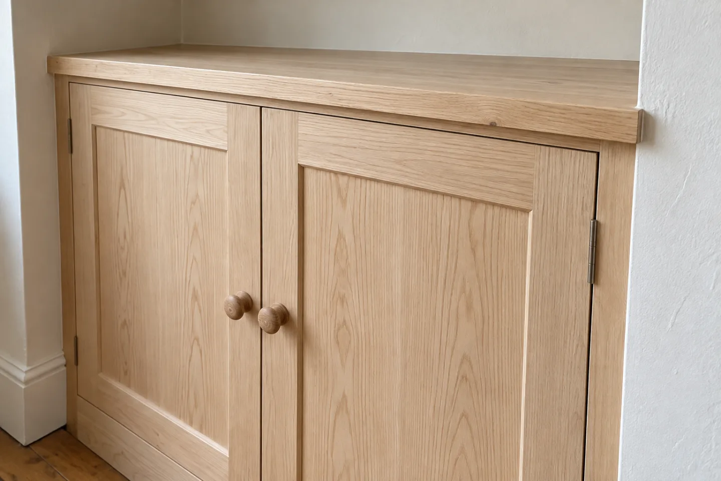 Close-up of unpainted cupboard doors, round timber knobs and the cupboard top beside a plaster wall.